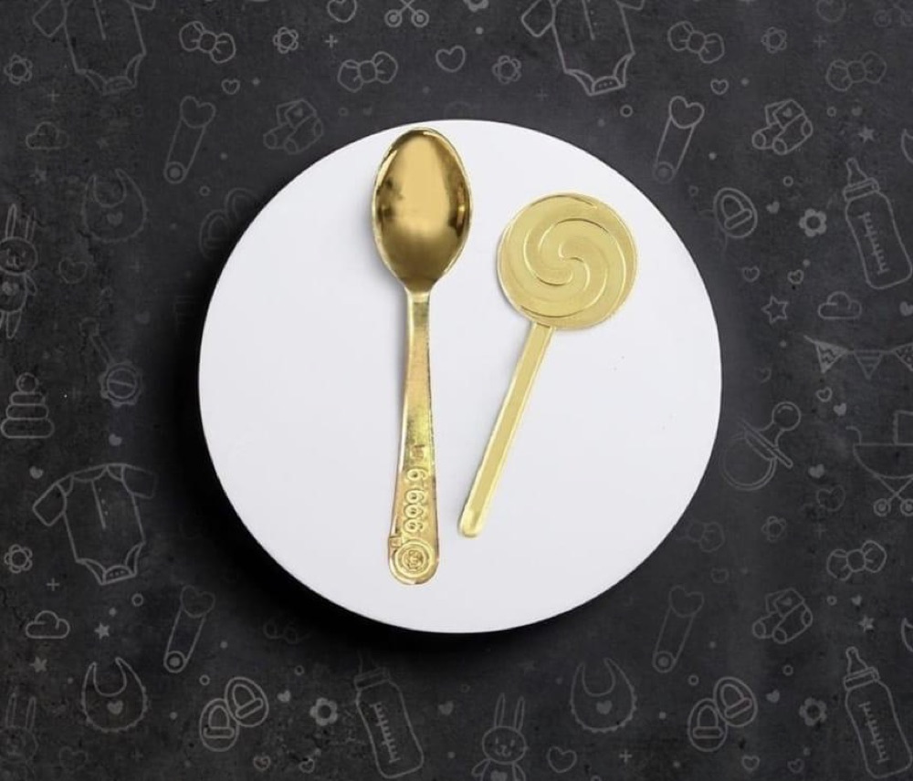 A gold baby spoon and a gold lollipop on a white plate